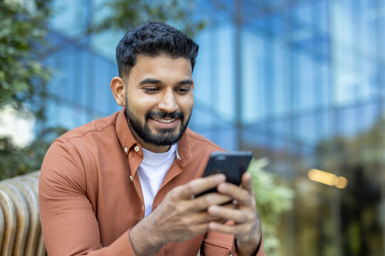 A smiling man with a beard looking at his phone while sitting outside near a modern building. - Powered by Adobe