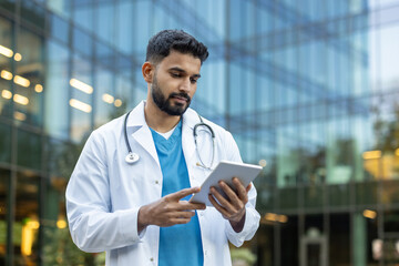 A doctor wearing a lab coat examines a tablet outside a modern medical building. He has a...