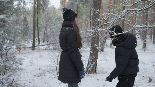 Son and mother playing on a winter walk in a snowy forest. The child plays a fun prank on his mom, shaking a tree branch. Snow from the tree falls, creating a snow shower for the happy family.