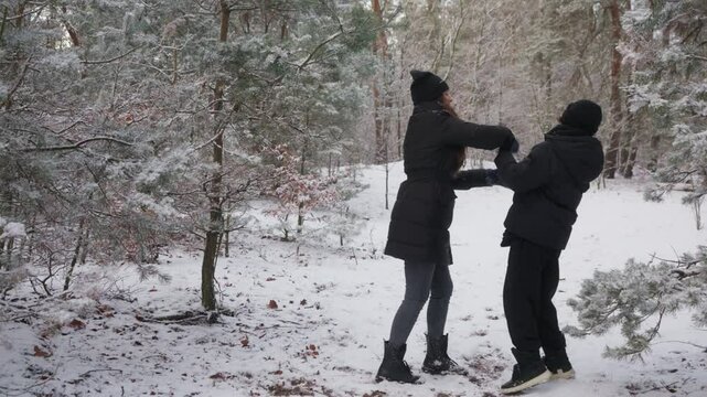 A mother plays a prank on her son in a winter forest, shaking snow on him from a tree. She then helps him brush the snow off his jacket collar. A playful family moment showing fun and care.