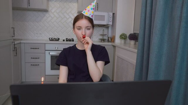 Teenage girl in party hat celebrating birthday online blowing horn during video call with piece of birthday cake and candle in front of laptop in kitchen at home.