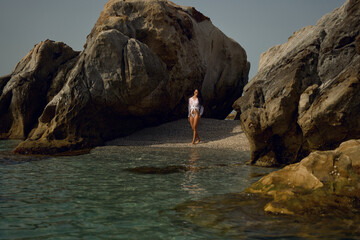 Young brunette woman in a soaked white shirt kneels near a rocky shore, posing seductively under...