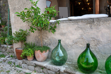 Several large, green, bulbous bottles stand on a low, old wall next to a house wall with some...