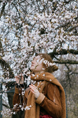 woman with red hair stands in front of a tree with pink flowers. She's wearing a brown coat, scarf.