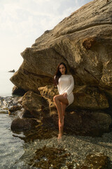 A slim brunette model in a short white crochet dress stands in shallow sea water, posing sensually with hands in hair during golden hour light.