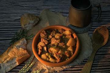 Rustic traditional stew with meat, potatoes and peas served in a clay bowl with bread and a vintage metal mug on a wooden table with natural warm light