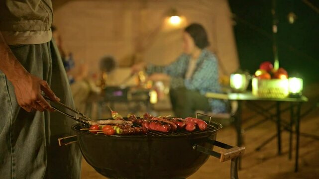 Man grilling sausages at campsite with his friend. Group of teenagers prepare dinner at campsite meal with grilled food and cozy lighting. Enjoying night picnic and barbecue in natural breeze.