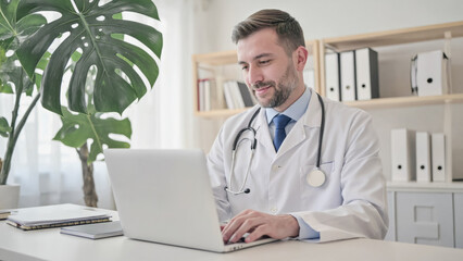 A young Caucasian man in a white lab coat works on a laptop in a modern office. A large green plant is visible in the background, along with shelves of files.