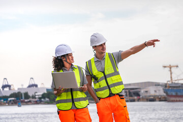 Two engineers wearing safety vests and helmets inspect port operations near the waterfront. They...