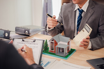 Real estate agent discusses and explains the steps of buying and selling a home. After that, they sign the sales contract in the office with a model home, laptop, calculator, documents, charts on the 