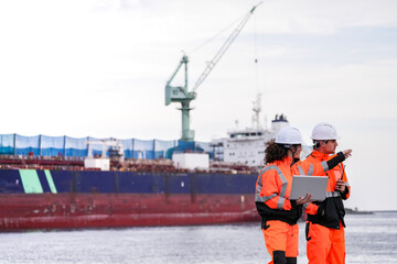 Two port engineers in high-visibility orange uniforms and hard hats communicate while inspecting...