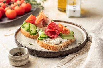 Delicious sandwiches with cream cheese, salami, cucumber, tomatoes on a plate on light background. Homemade breakfast