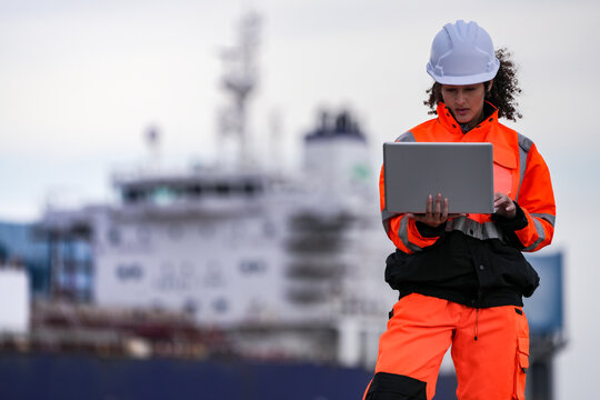 Two port engineers in high-visibility orange uniforms and hard hats communicate while inspecting dockside logistics using a laptop and walkie-talkie, symbolizing teamwork and safety. - Powered by Adobe