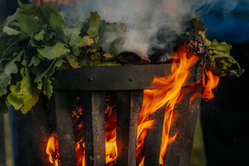 A metal fire basket with tall, bright orange flames burns on a grassy lawn, with embers rising into...