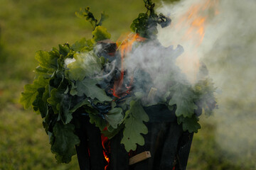 A metal fire basket with tall, bright orange flames burns on a grassy lawn, with embers rising into...