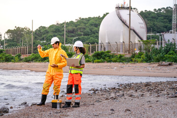 Two environmental engineers in safety gear work at a shoreline near gas storage tanks. One collects...