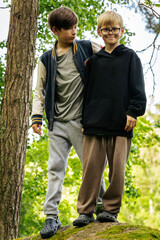 Two boys stand together on a mossy rock in a forest, dressed in casual clothes and smiling amid...