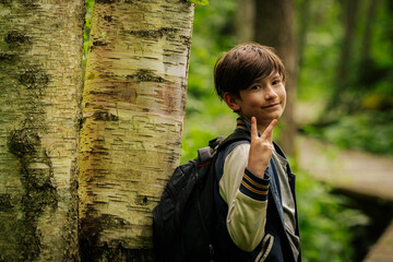 Smiling boy with a backpack leaning against a birch tree on a forest boardwalk, surrounded by lush...