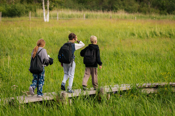 Three kids walk along a narrow wooden path through tall green grass in a peaceful field, enjoying a...