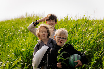 Three children stand in tall green grass making playful expressions, with one flashing a peace sign and others smiling and goofing around in natural light.