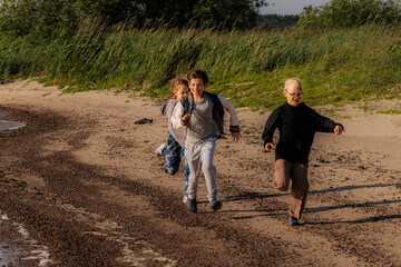 Fototapeta premium Two children walk and run along a sandy shoreline toward a friend standing near tall grass, with gentle waves lapping the beach under a bright sky.