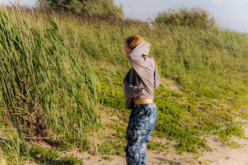 A young girl walks playfully along a sandy beach with wind blowing her hair, dressed in a gray hoodie and patterned pants, near tall green grass.