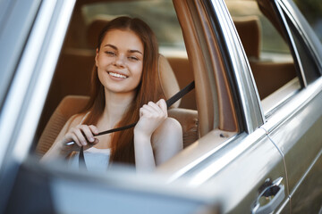 Young woman smiling while fastening her seatbelt in a car, expressing happiness and safety, dressed casually in a light top, with a warm, outdoor background.