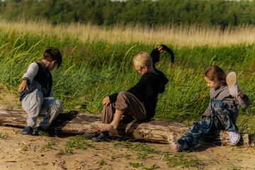 Three children sit and play on a driftwood log at the edge of a grassy dune, enjoying a sunny and...