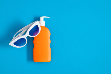 Sunscreen. Stylish white glasses and orange bottle of sunscreen lotion on blue background