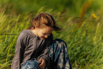 A smiling girl sits on a weathered log in a grassy field, wearing star-patterned pants and a gray...