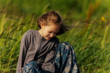 A smiling girl sits on a weathered log in a grassy field, wearing star-patterned pants and a gray...