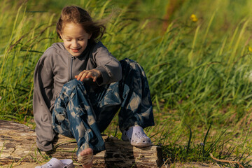 A smiling girl sits on a weathered log in a grassy field, wearing star-patterned pants and a gray...