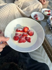 Close-up of a hand holding a bowl of fresh strawberries topped with sugar and cream, enjoyed outdoors in cozy surroundings.