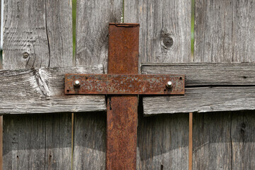 Rusty metal cross attached to a weathered wooden fence. The texture of the wood is rough and aged, showcasing natural imperfections and a rustic appearance.