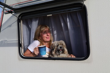 A woman with a cup in her hand and her dog look out through the open window of a camper van,...
