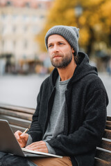 Thoughtful entrepreneur working remotely on a city bench with laptop and notebook