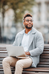Thoughtful entrepreneur working remotely on a city bench with laptop and notebook