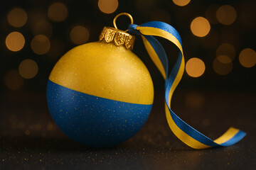 Christmas bauble in Ukrainian flag colors with golden glitter and matching ribbon on dark background. Festive, patriotic holiday decoration concept