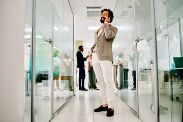 Smiling businessman talking on mobile phone in modern office corridor with colleagues discussing in...