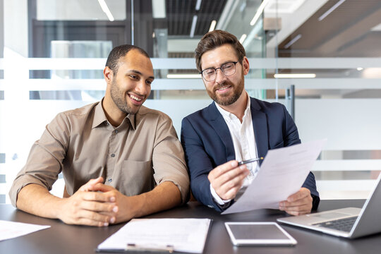 Two businessmen are reviewing documents in a modern office, discussing plans, and smiling. Collaboration and business are key themes.