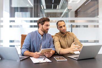 Two diverse businessmen collaborate in a modern office, discussing project details with laptops and...