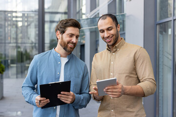 Two diverse businessmen review a tablet, possibly discussing a project or presenting ideas, smiling in a modern outdoor setting.