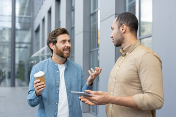 Two businessmen are engaged in a discussion outdoors, with one holding a coffee cup and the other a tablet.