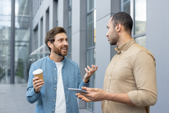 Two businessmen are engaged in a discussion outdoors, with one holding a coffee cup and the other a tablet. - Powered by Adobe