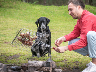 Man grilling meat outdoors near fire pit while dog watches hungrily, summer picnic, outdoor cooking, friendship with pet.