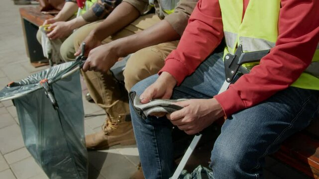 Unrecognizable male and female volunteers holding plastic bag with trash and gloves while resting on bench