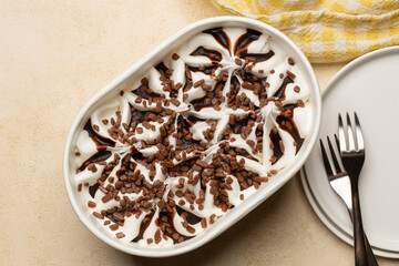 Vanilla ice cream with chocolate sauce and chocolate pieces in a paper box on a beige table, plates and forks. Food background.