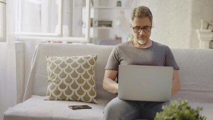 Older man working online with laptop computer at home sitting on couch in living room. Home office, browsing internet. Portrait of happy, mature age, middle age, mid adult man in 50s, smiling. - Powered by Adobe