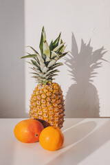 Sunny pineapple stands tall beside a mango and orange, casting dramatic shadows against a stark white wall in a warm afternoon light