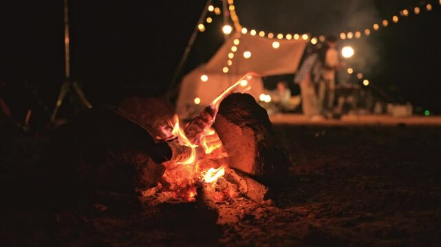 Close-up of rustic campfire burning in outdoor night setting glowing campfire at night with tent and string lights in background, Relaxing camping moment beside campfire in wilderness